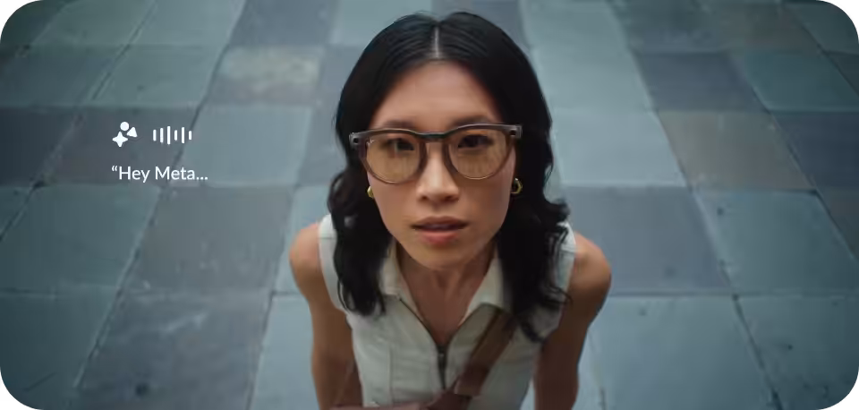 "A woman with long black hair, wearing sunglasses and a sleeveless white top, looks up toward the camera while standing on a tiled outdoor surface. A speech bubble with 'Hey Meta...' and sound wave icons appear beside her, suggesting interaction with a virtual assistant from her glasses."