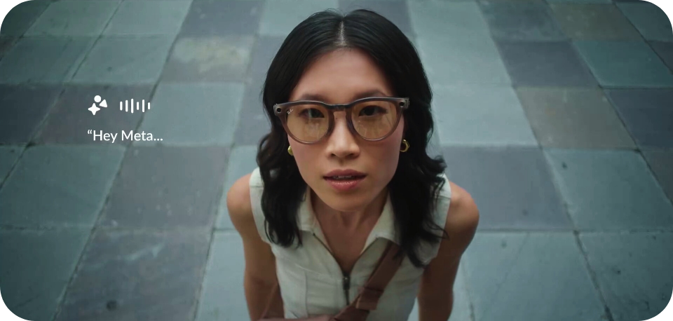 "A woman with long black hair, wearing sunglasses and a sleeveless white top, looks up toward the camera while standing on a tiled outdoor surface. A speech bubble with 'Hey Meta...' and sound wave icons appear beside her, suggesting interaction with a virtual assistant from her glasses."