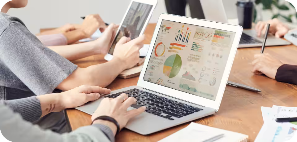 A group of professionals collaborating at a wooden desk, with a laptop displaying colorful charts and graphs, representing data analysis and teamwork in a modern office setting.