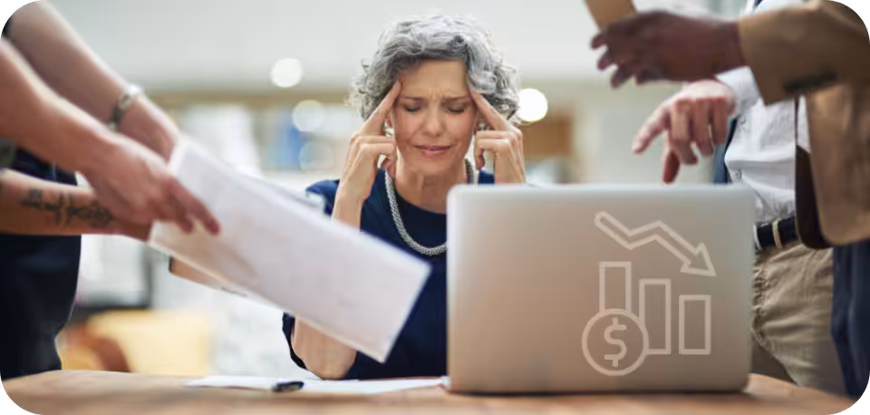 Stressed businesswoman at a desk surrounded by colleagues showing documents and mobile devices, with a laptop displaying a declining financial graph, representing the downsides of paid social media campaigns.