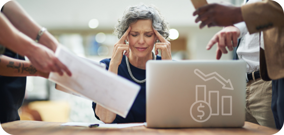 Stressed businesswoman at a desk surrounded by colleagues showing documents and mobile devices, with a laptop displaying a declining financial graph, representing the downsides of paid social media campaigns.